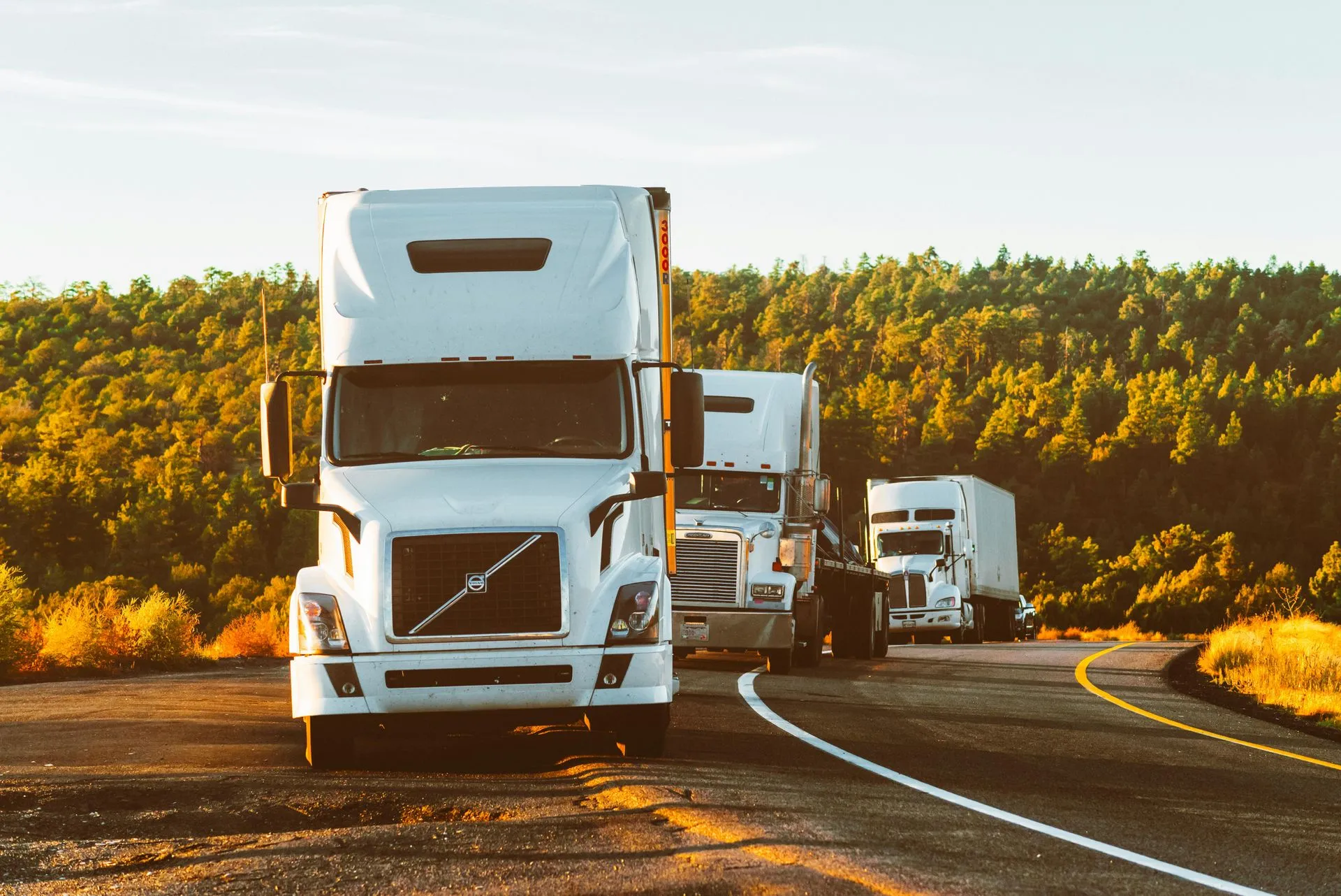 Three white semi-trucks driving along a curving highway